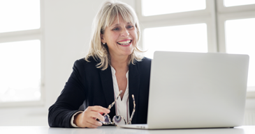 A woman in business attire sitting at a desk smiling while looking at an open laptop.