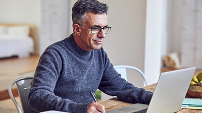 A man in a sweater sitting at a wooden table, using his laptop while taking notes 