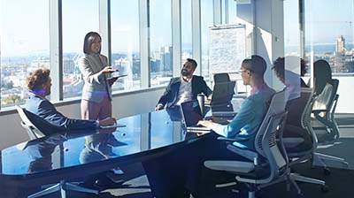 Group of six people in conference room listening to a woman speak with a presentation board