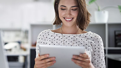 A woman in a black and white polka dot blouse smiling down at a tablet that she is holding with both hands 