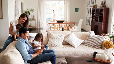 A family sitting on a sectional in their living room. The dad is sitting and holding his daughter while looking at a tablet together. The mother is holding her son while leaning over the couch behind the dad. 