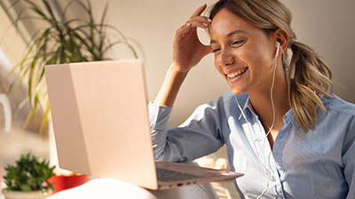 A woman wearing earphones while laughing at something on her laptop. Her right hand is touching her forehead.