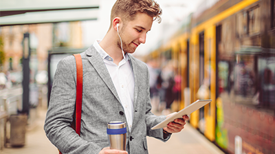 A man in a gray blazer wearing headphones and looking down at a tablet in one hand, while holding a travel mug in his other hand