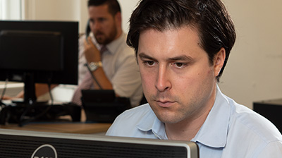 Man dressed in business casual attire looking at computer screen while a colleague in the background holds phone and uses computer.