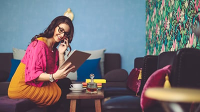 Smiling woman in brightly colored clothing, holding a cell phone to her ear while looking down at a tablet device in her left hand. She’s sitting on a couch with a coffee table in front of her.