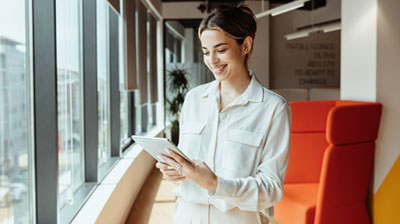 A woman smiling at a tablet while standing in front of a wall of windows with an orange chair in the background.