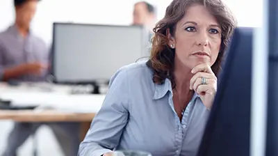 Woman in business casual attire sitting at desk using a desktop computer and smiling.