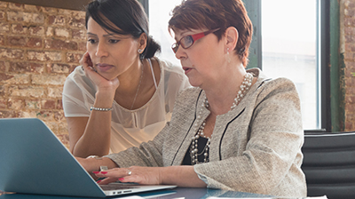 Two women in casual attire looking down at an open laptop in front of them. One woman is standing next to the other who is sitting.