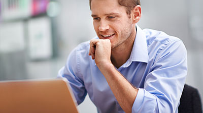 Man in business casual attire sitting and smiling while using a laptop.