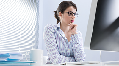 Woman in business attire sitting at desk and staring at the computer with hand to her chin 