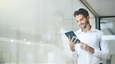 Man dressed in business casual attire leaning on window smiling and using tablet.