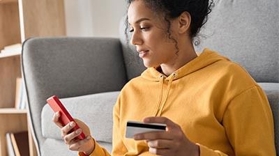 A woman in an orange hoodie using a red smart phone while holding a credit card with her other hand