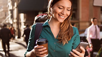 Woman dressed in casual attire walking down the street holding a disposable coffee cup and looking at her smartphone with backpack on her back.