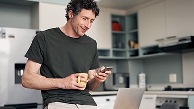 A man standing in his kitchen, holding a yellow mug while using his smart phone